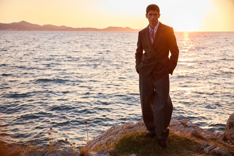 A man in a suit poses on a rocky outcrop at sunset with the ocean and distant mountains in the background, creating a stylish and scenic composition.