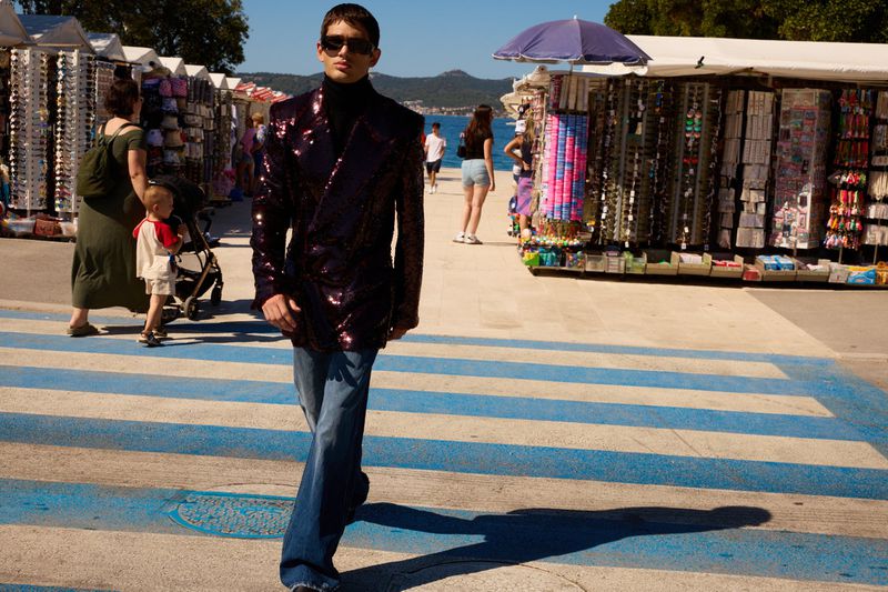 Man in sunglasses and sequin blazer walks on a blue crosswalk near vendor stalls and the ocean on a sunny day by the coast.