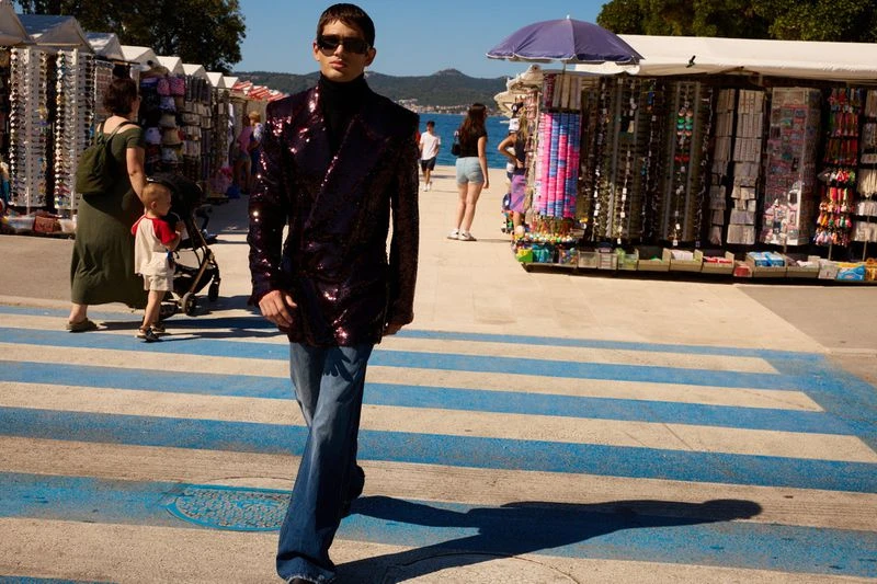 Man in sunglasses and sequin blazer walks on a blue crosswalk near vendor stalls and the ocean on a sunny day by the coast.