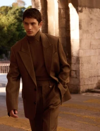 Model Luka Tesic wearing a brown tailored suit and turtleneck walking outdoors near stone architecture