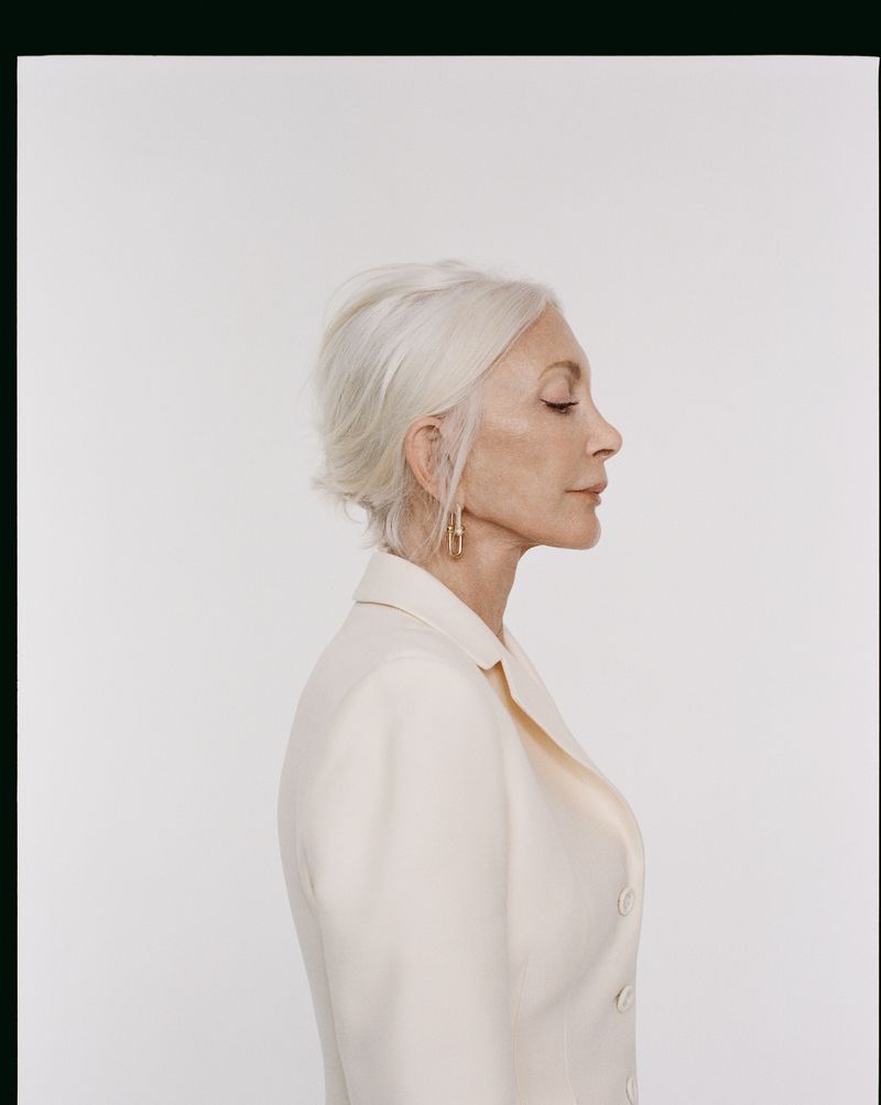 Profile view of an elderly woman with short white hair in a cream-colored jacket, wearing gold earrings against a white backdrop.