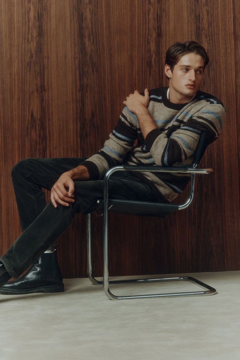 A young man in a striped sweater sitting in a chrome chair against a wood paneled wall wearing dark denim and shiny black leather boots.
