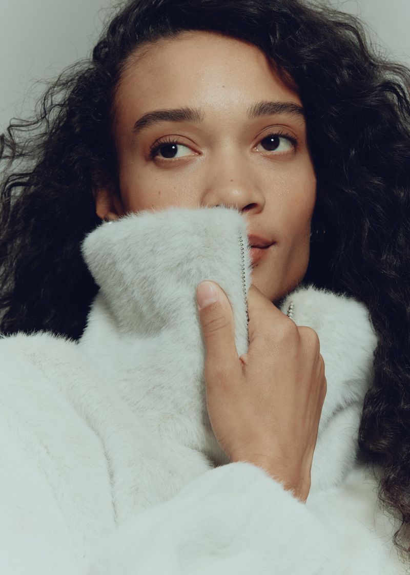 Close-up studio shot of a woman with curly hair in a white jacket with a high collar, partially obscuring her face with the collar.