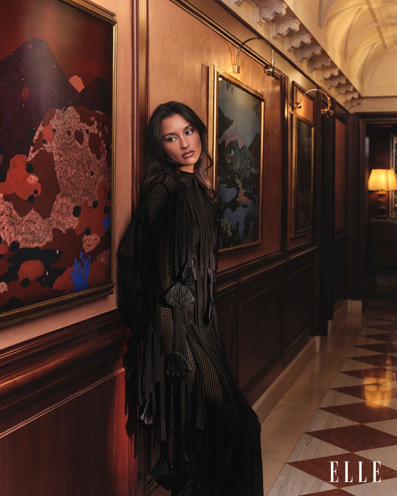 Portrait of a woman leaning against a wood-paneled wall, wearing a black net dress with fringe accents, in a hallway with artwork.