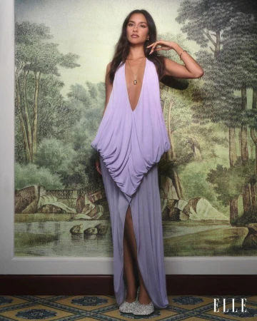 A fashionable woman posing in a lilac gown in front of a painted backdrop, wearing sparkling shoes and jewelry, and the Elle logo.