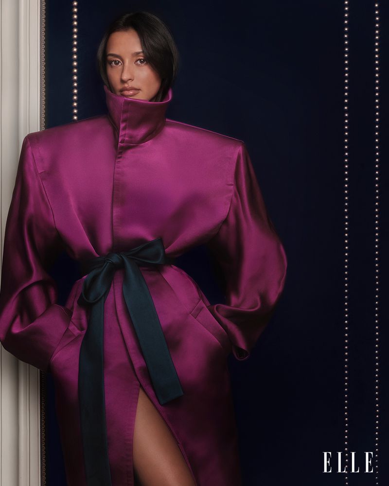 Portrait of an adult woman in a purple, shiny coat cinched with a dark bow, posing in front of a dark wall with light trim.
