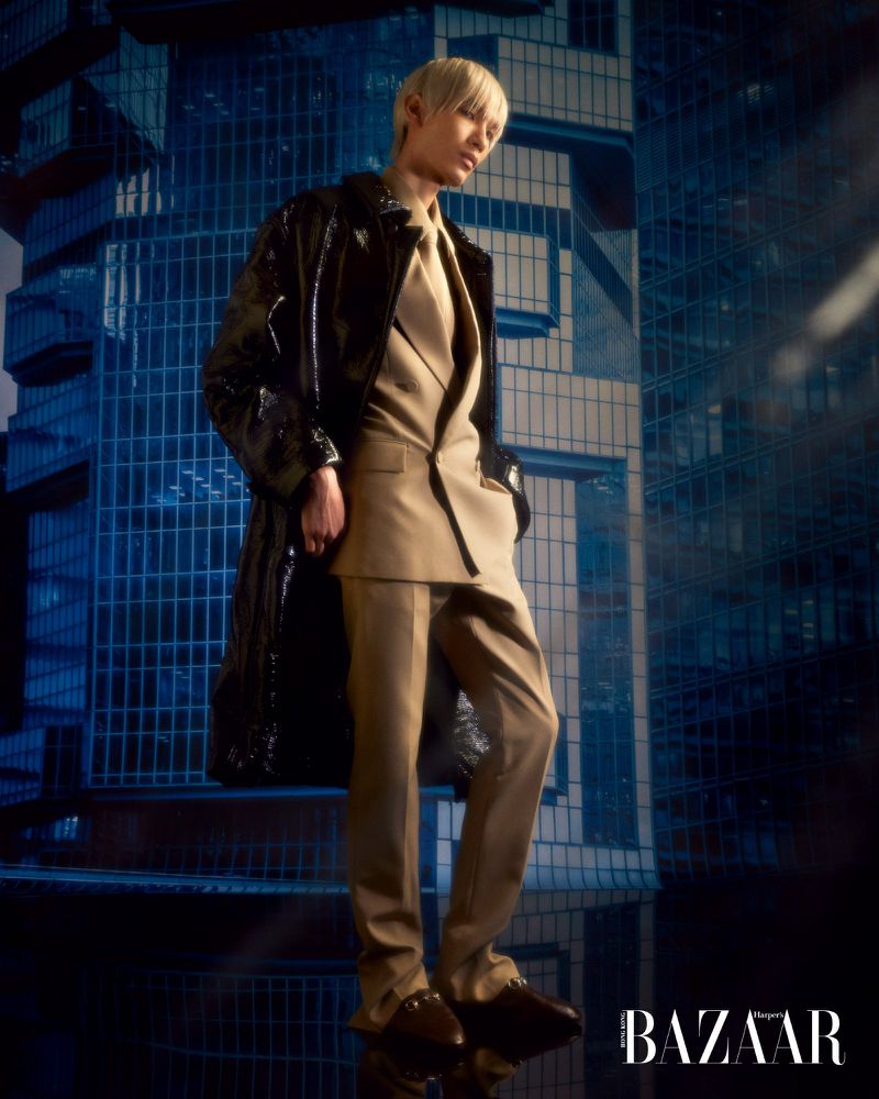 Fashion editorial shot of a male model wearing a tan suit and black patent leather coat, posing against a dark blue building backdrop.