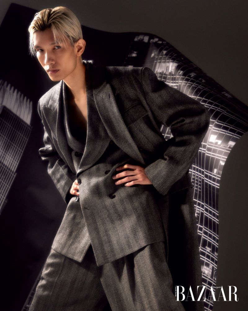 An adult male model poses in a grey suit in front of a grey background with an image of high rise buildings; he is staring forward.