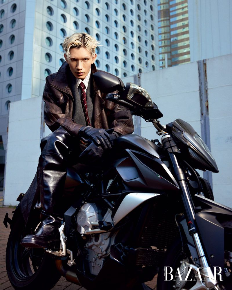 An adult male with bleached-blond hair poses on a dark motorcycle with a modern building in the background wearing a coat and gloves.