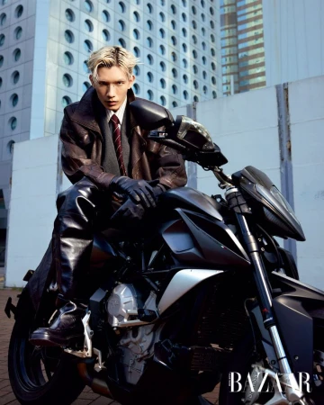 An adult male with bleached-blond hair poses on a dark motorcycle with a modern building in the background wearing a coat and gloves.