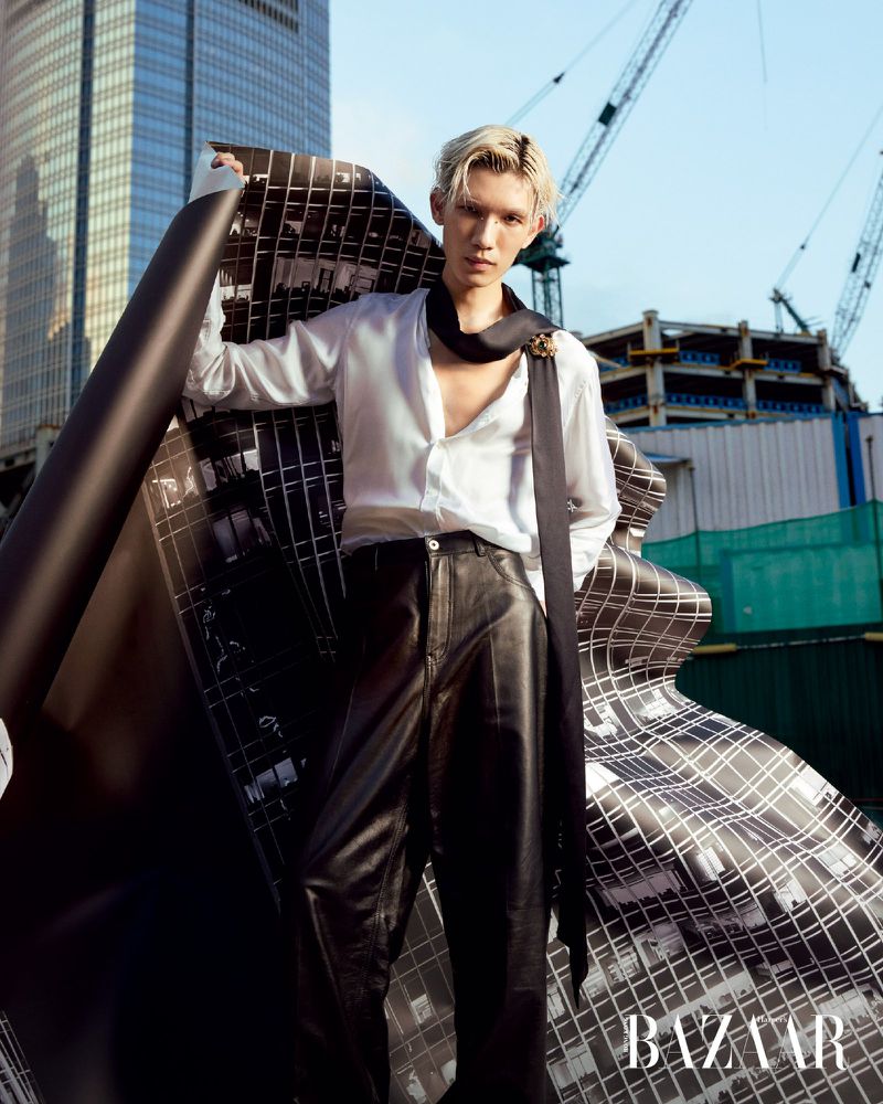 Fashionable male model in unbuttoned white shirt and leather pants, posing with a printed textile against a city construction backdrop.