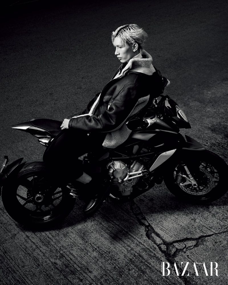 Monochrome shot of an adult man with bleached hair in a jacket sitting on a black motorcycle on cracked pavement, fashion editorial style.