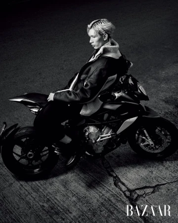 Monochrome shot of an adult man with bleached hair in a jacket sitting on a black motorcycle on cracked pavement, fashion editorial style.