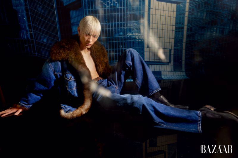 Fashion editorial shot of a blonde adult male model posing on a platform in a denim jacket with a fur collar and blue denim pants.