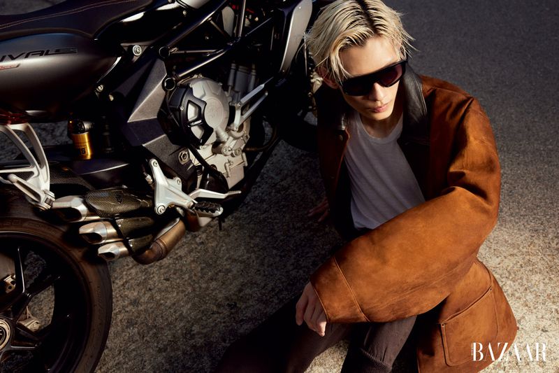 Man in a brown coat sits next to a black motorcycle, wearing sunglasses and a white t-shirt, with the Bazaar logo visible.
