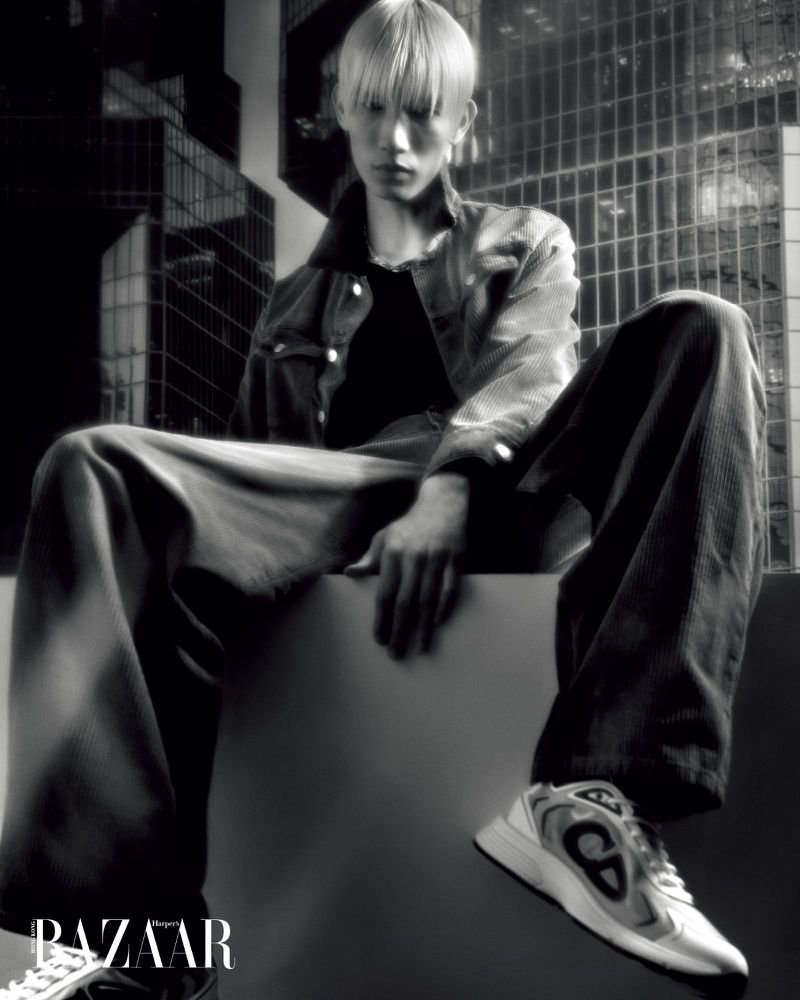 Black-and-white fashion shot of a young adult with blonde hair in a jacket and pants, sitting on a cube against a city backdrop.