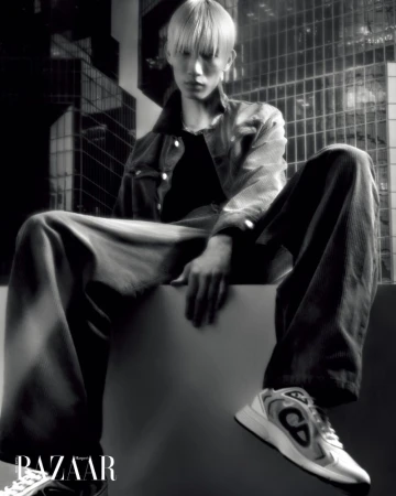 Black-and-white fashion shot of a young adult with blonde hair in a jacket and pants, sitting on a cube against a city backdrop.