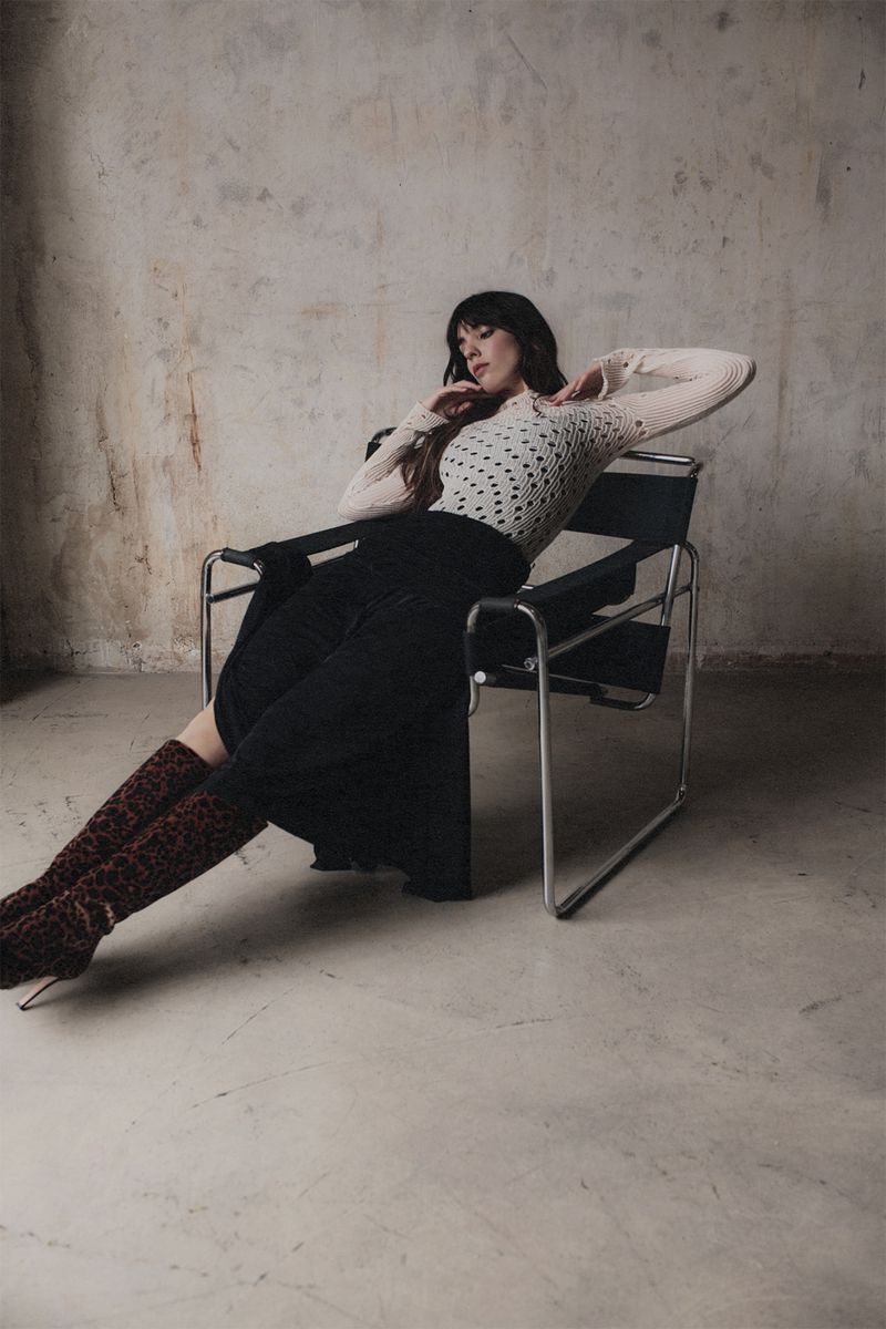 A woman with dark hair sits in a black chair with legs crossed and arms raised, wearing a sheer white top and long black skirt in a studio.
