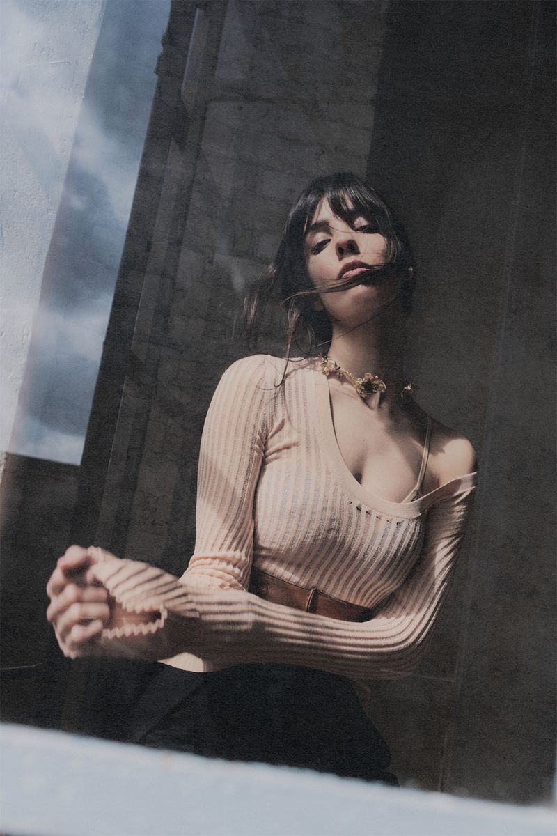 Woman with dark hair poses in front of a tall stone structure on a sunny day, wearing a fitted light-peach top and dark pants.