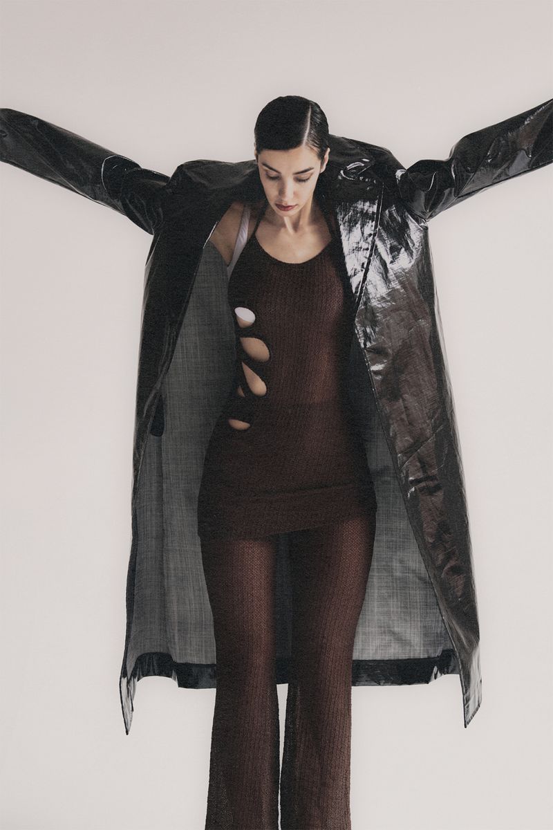 A woman stands in a studio wearing a brown dress with cutouts and a black, shiny coat, her arms slightly outstretched against a white background.