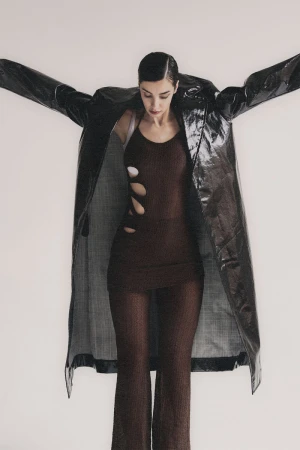 A woman stands in a studio wearing a brown dress with cutouts and a black, shiny coat, her arms slightly outstretched against a white background.