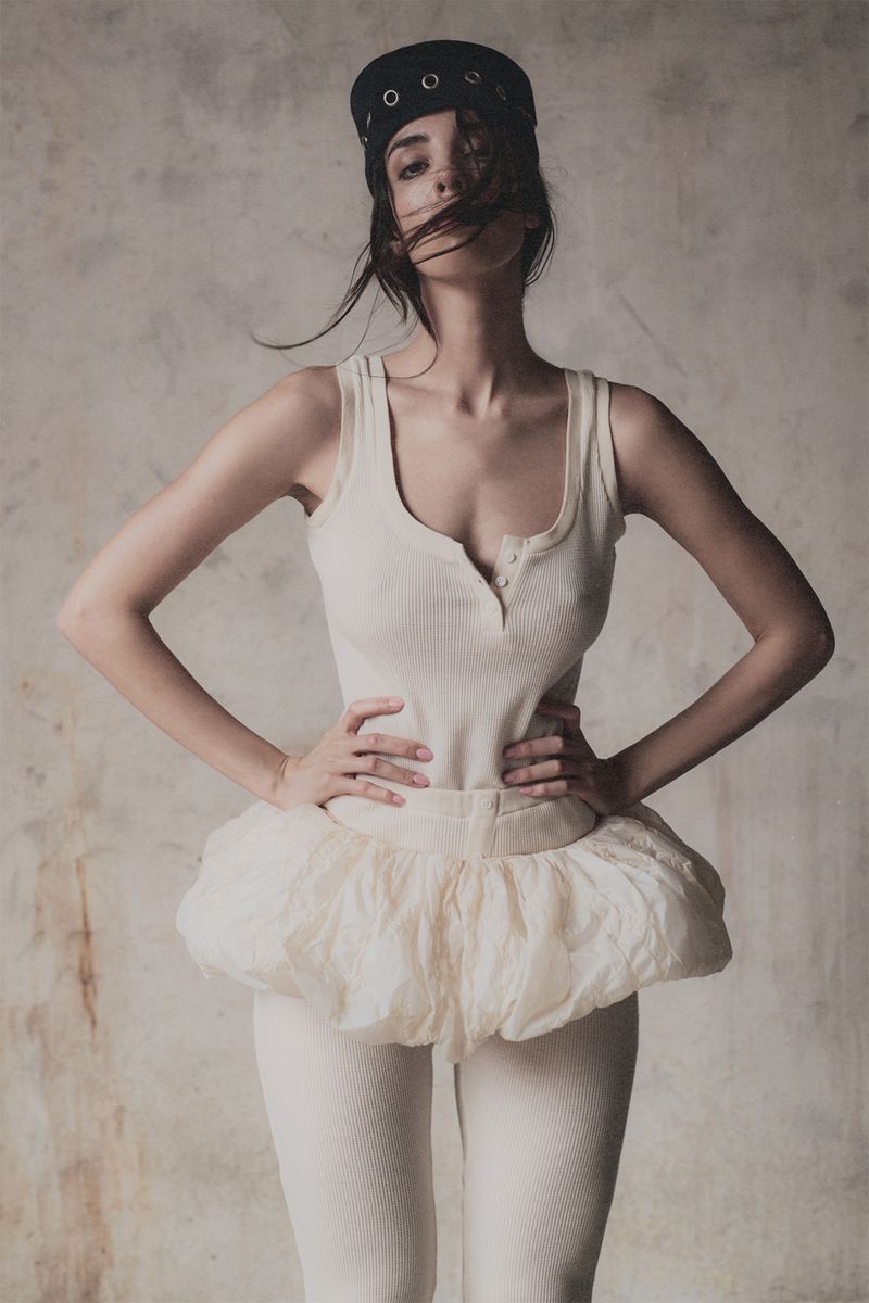 A young woman poses indoors wearing a tank top, tutu, leggings, and hat. The lighting is soft, and the background is a textured wall.