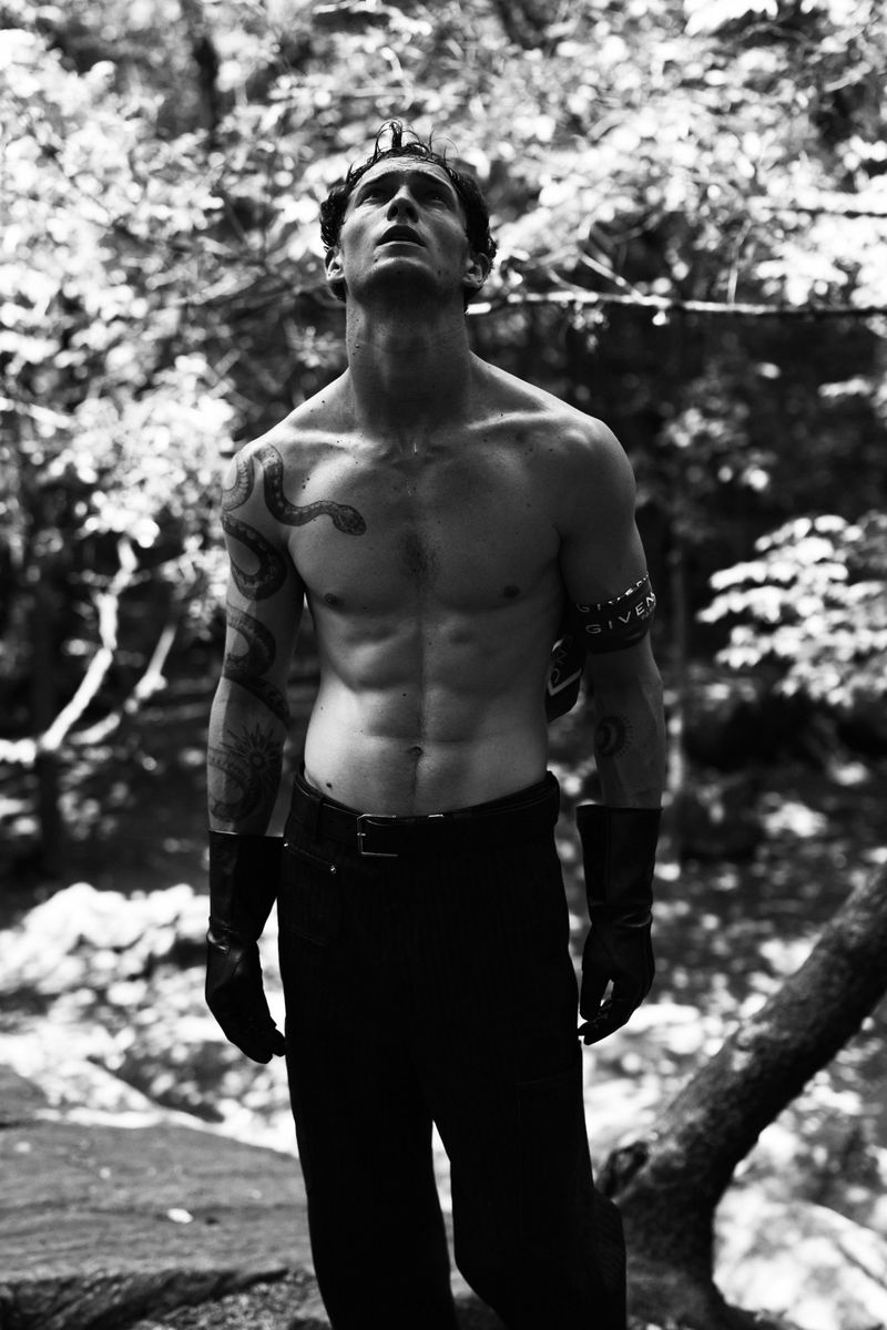 Black and white portrait of a muscular adult male model standing shirtless in a forest setting, looking up, and wearing black pants.