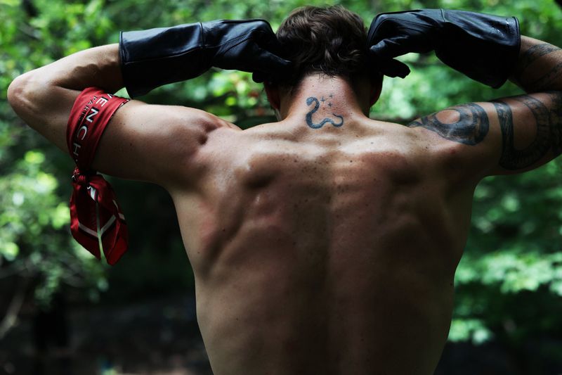 Shirtless man seen from behind wearing long black leather gloves and a red printed armband in lush greenery.