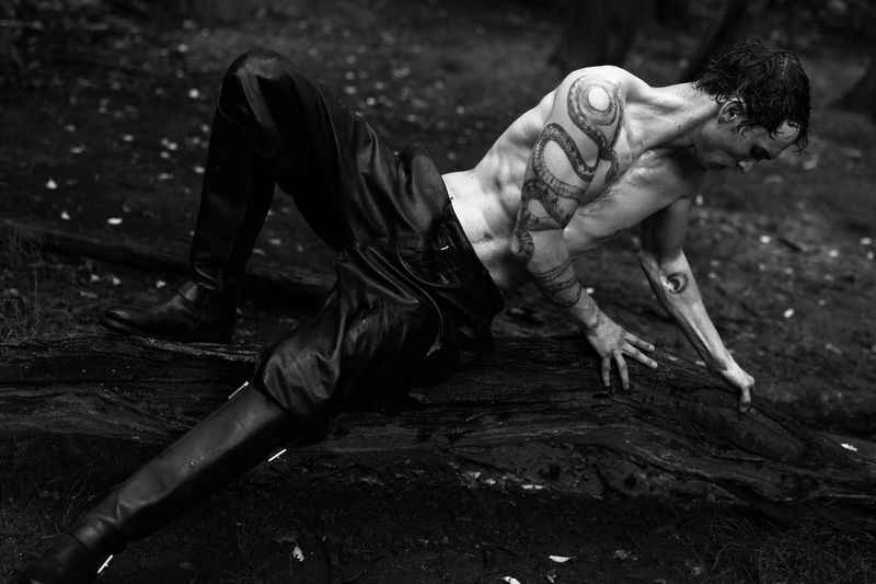 Adult male model poses shirtless with snake tattoo in black leather pants and boots, outdoors in a black and white fashion photo.