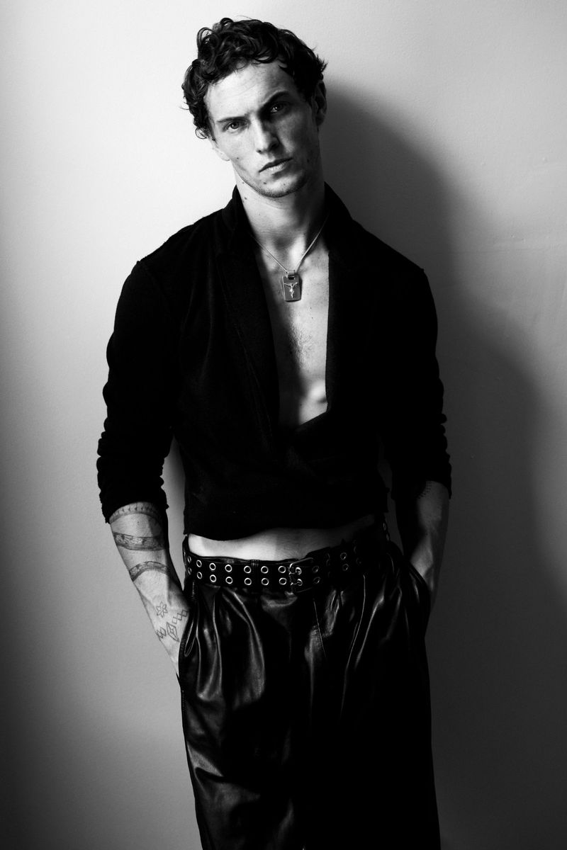 Black and white studio portrait of a slender adult male model wearing a cropped shirt and leather pants with his hands in his pockets.