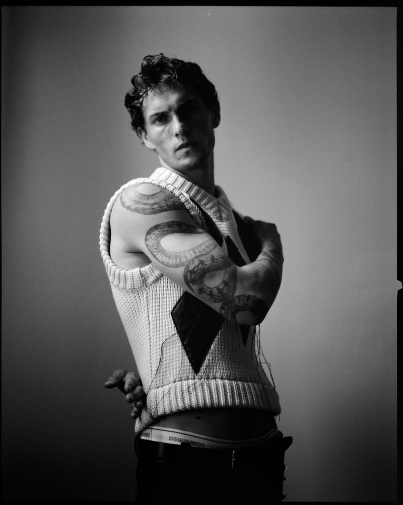 Black and white portrait of a young adult male with snake tattoos, wearing a sweater vest, standing indoors against a plain backdrop.