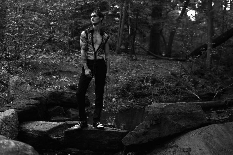 Black and white portrait of a male fashion model with tattoos standing on rocks in a forest wearing open vest and black pants.