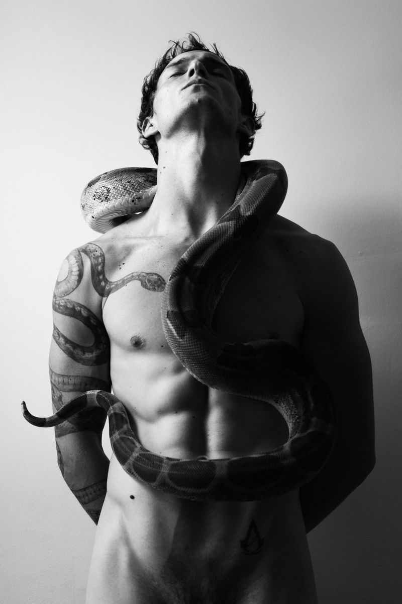 Striking black and white portrait of a shirtless man with tattoos and a snake wrapped around his body, eyes closed, against a plain background.