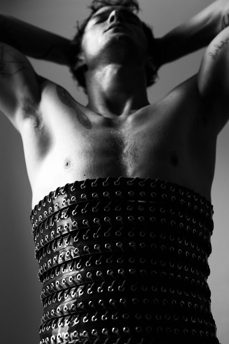 Black and white portrait of a muscular adult man with hands behind his head, wearing a layered leather-like corset, looking up in an indoor setting.