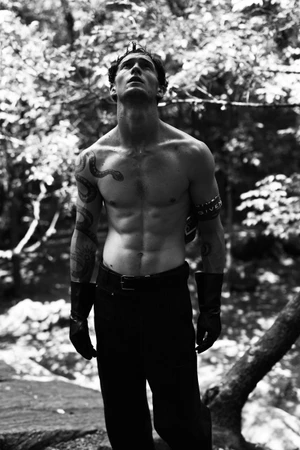 Black and white portrait of a shirtless man with arm tattoos and black leather gloves standing outdoors in dappled sunlight.