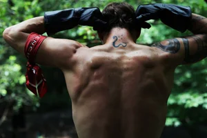 Shirtless man seen from behind wearing long black leather gloves and a red printed armband in lush greenery.