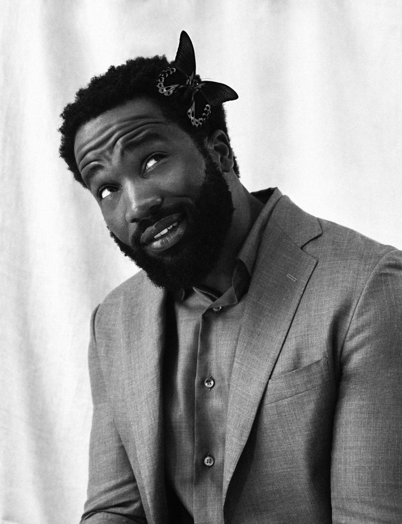 Monochromatic portrait of an African American man in a suit with a butterfly hair clip looking up against a draped white background.