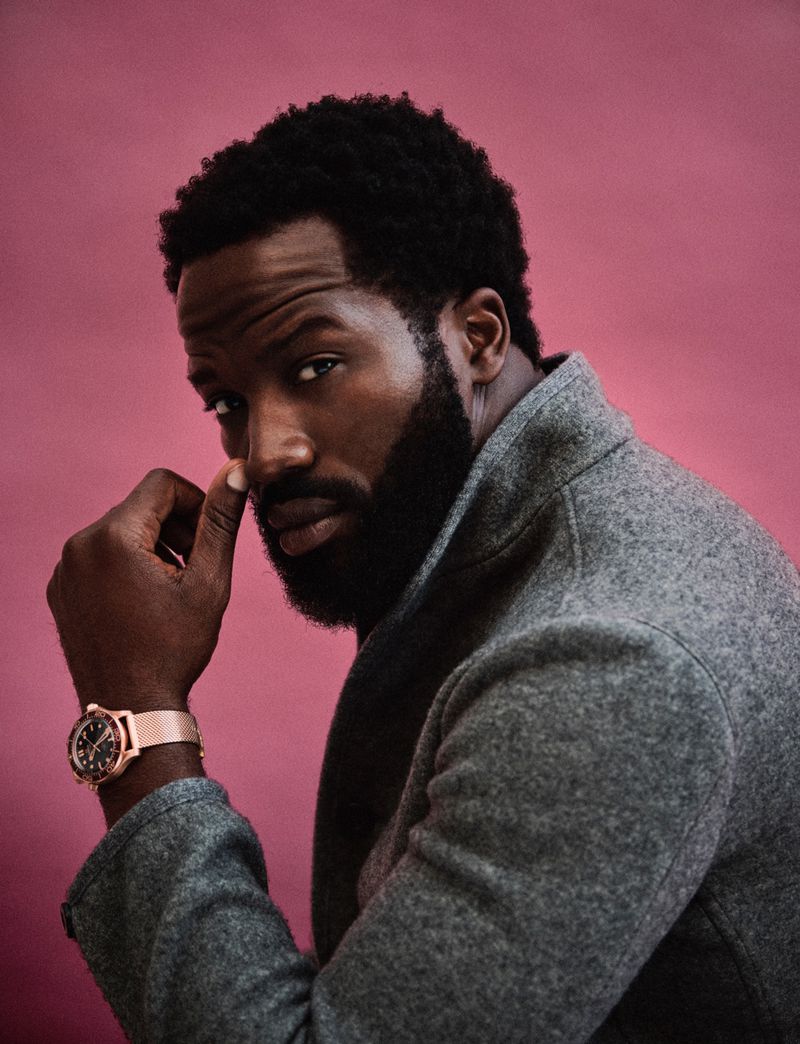 Portrait of an adult Black man in a grey coat against a pink background, looking at the camera with a slight smile and wearing a watch.