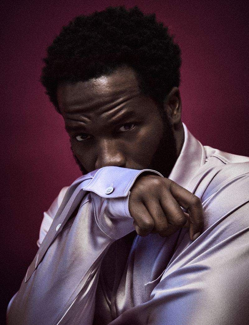 Portrait of handsome adult Black man with dark hair and light purple satin shirt against dark rose background in a close-up studio shot.