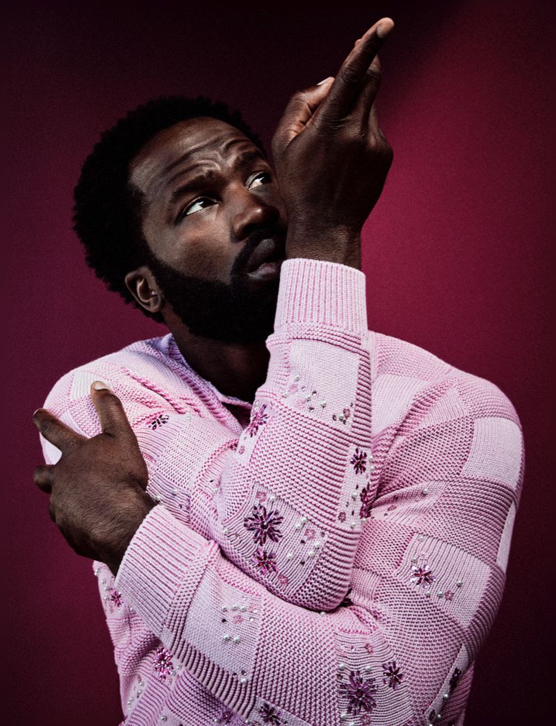Portrait of a dark-skinned man with a beard in a pink patterned sweater pointing up with a serious expression against a red backdrop.