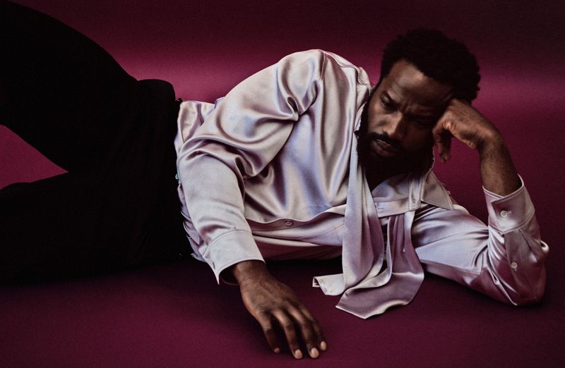 Portrait of a Black man reclining in a satin shirt on a purple surface, with focus on his expression and the shirt's draped collar.