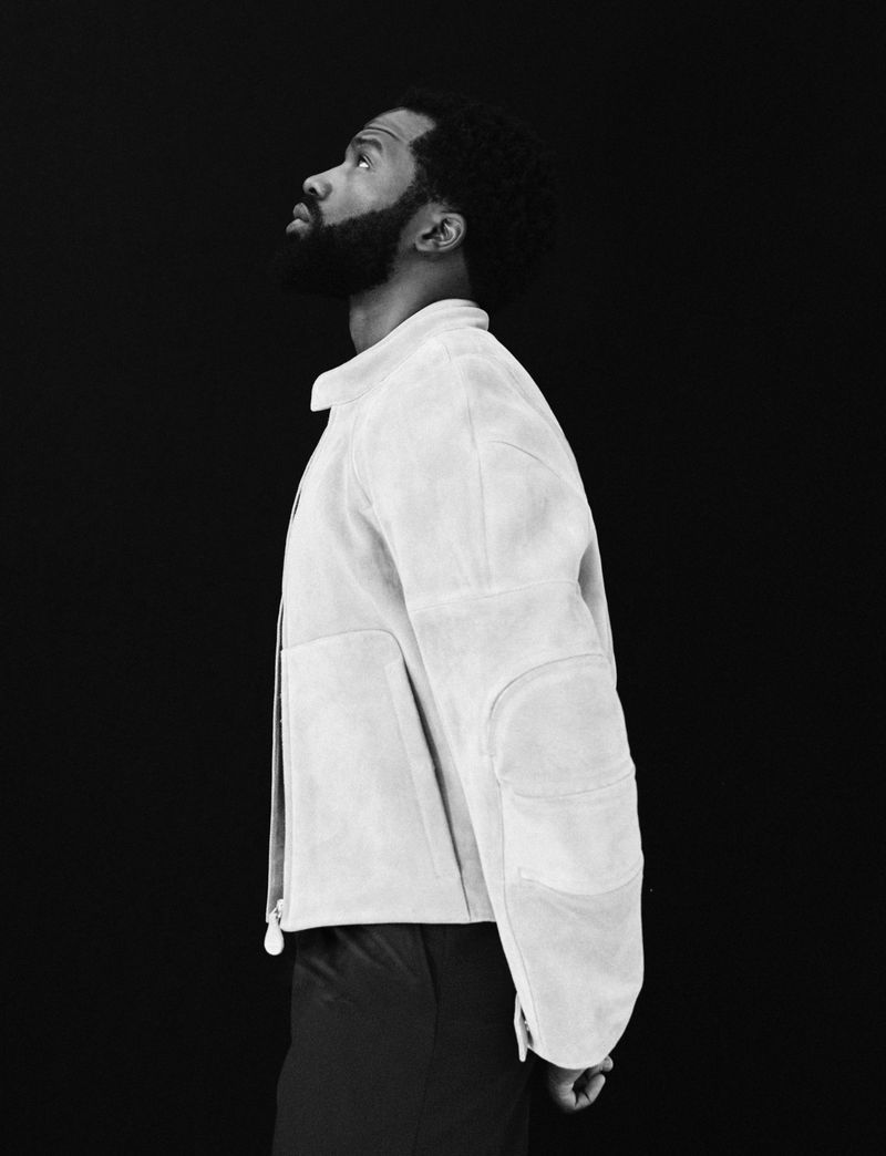 Profile shot of an adult man with a beard looking upward, wearing a jacket, against a dark background in a studio setting.