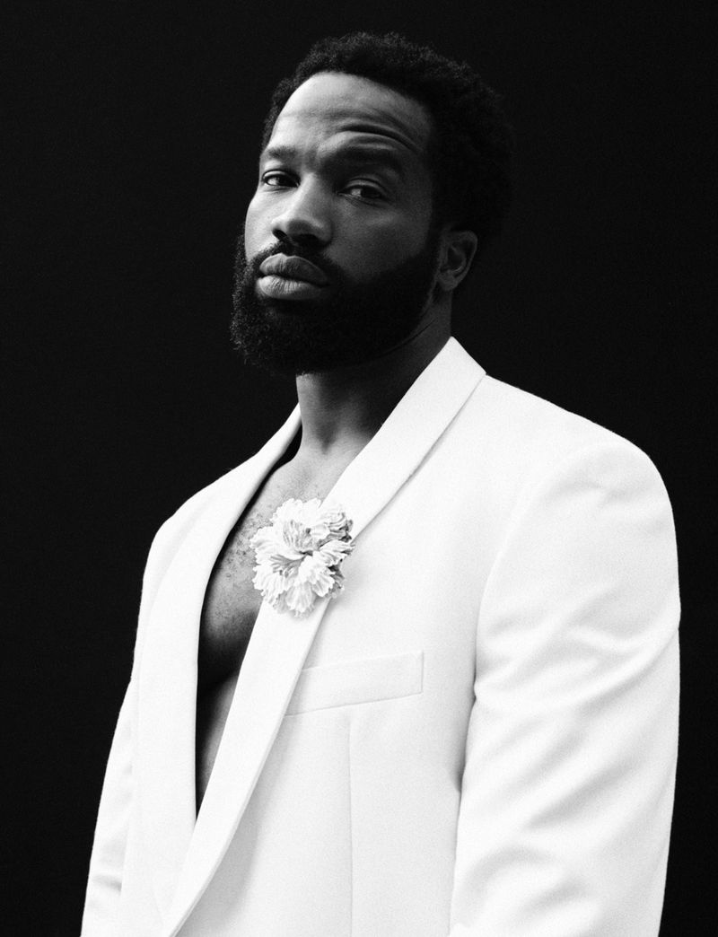 Portrait of a bearded Black male model in a white jacket with a flower against a black background in a photo studio setting.