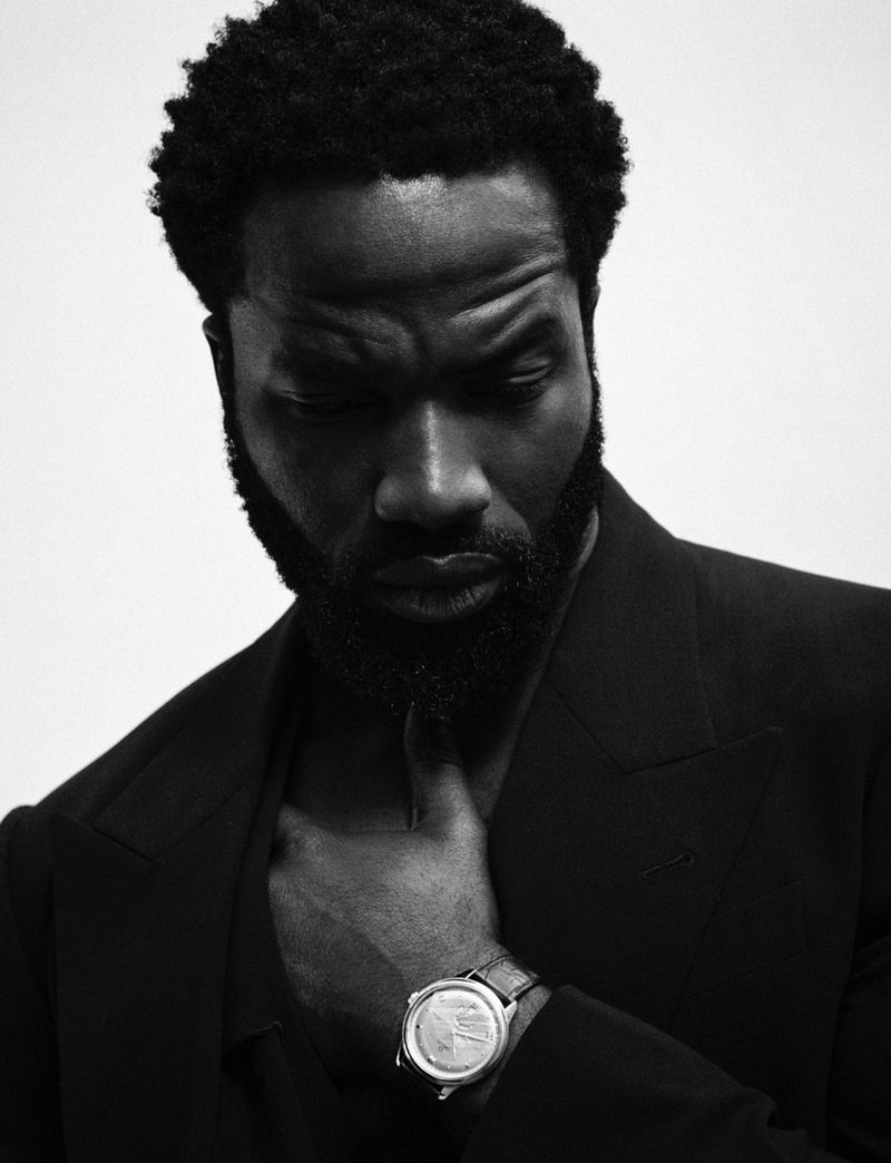 Monochrome portrait of a Black man in a suit with a beard, looking down, hand at his neck, and wearing a wristwatch against a white backdrop.
