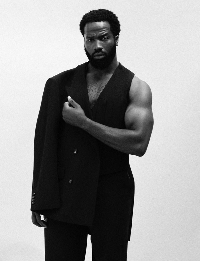 Portrait of an African-American male model in a black suit vest with the jacket draped over his shoulder against a white backdrop.
