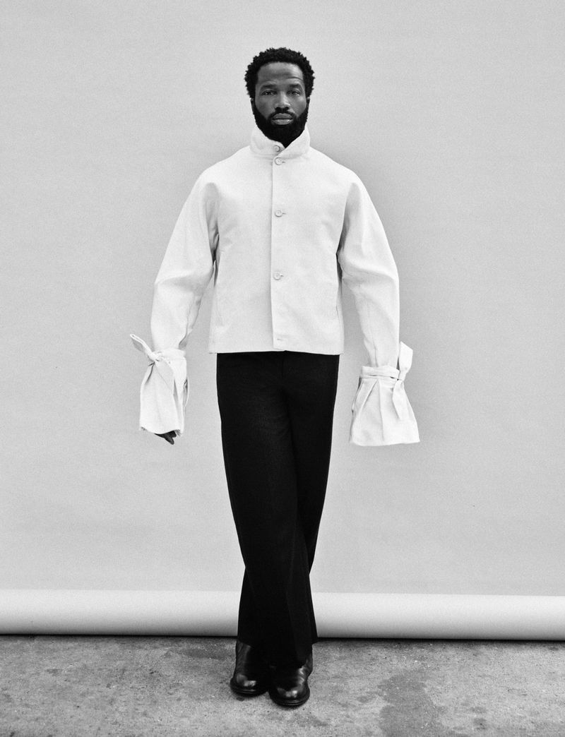 Black and white studio shot of adult male in white button-down shirt with wide sleeves and black pants, standing on concrete surface.