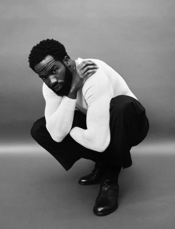 Black and white portrait of an African American male in casual attire, kneeling indoors against a neutral backdrop in photo studio lighting.