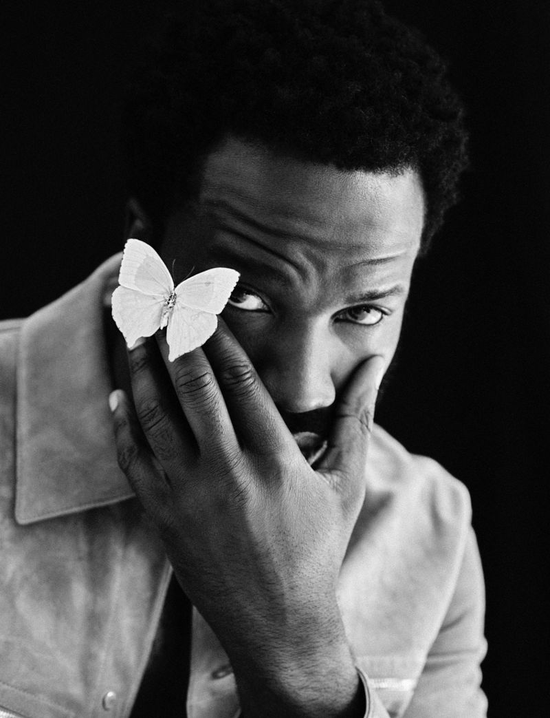 Portrait of a Black man with a butterfly on his hand, covering part of his face, and wearing a light colored jacket against a dark background.