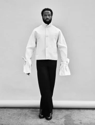 Black and white portrait of actor Sope Dirisu wearing a light jacket with tied cuffs and dark trousers against a plain background