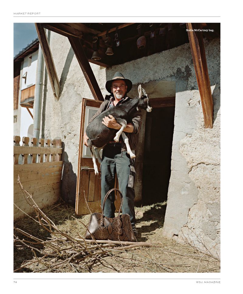 Man holding a goat near a barn with a designer bag on the ground; rustic outdoor scene with natural light and rural architecture.